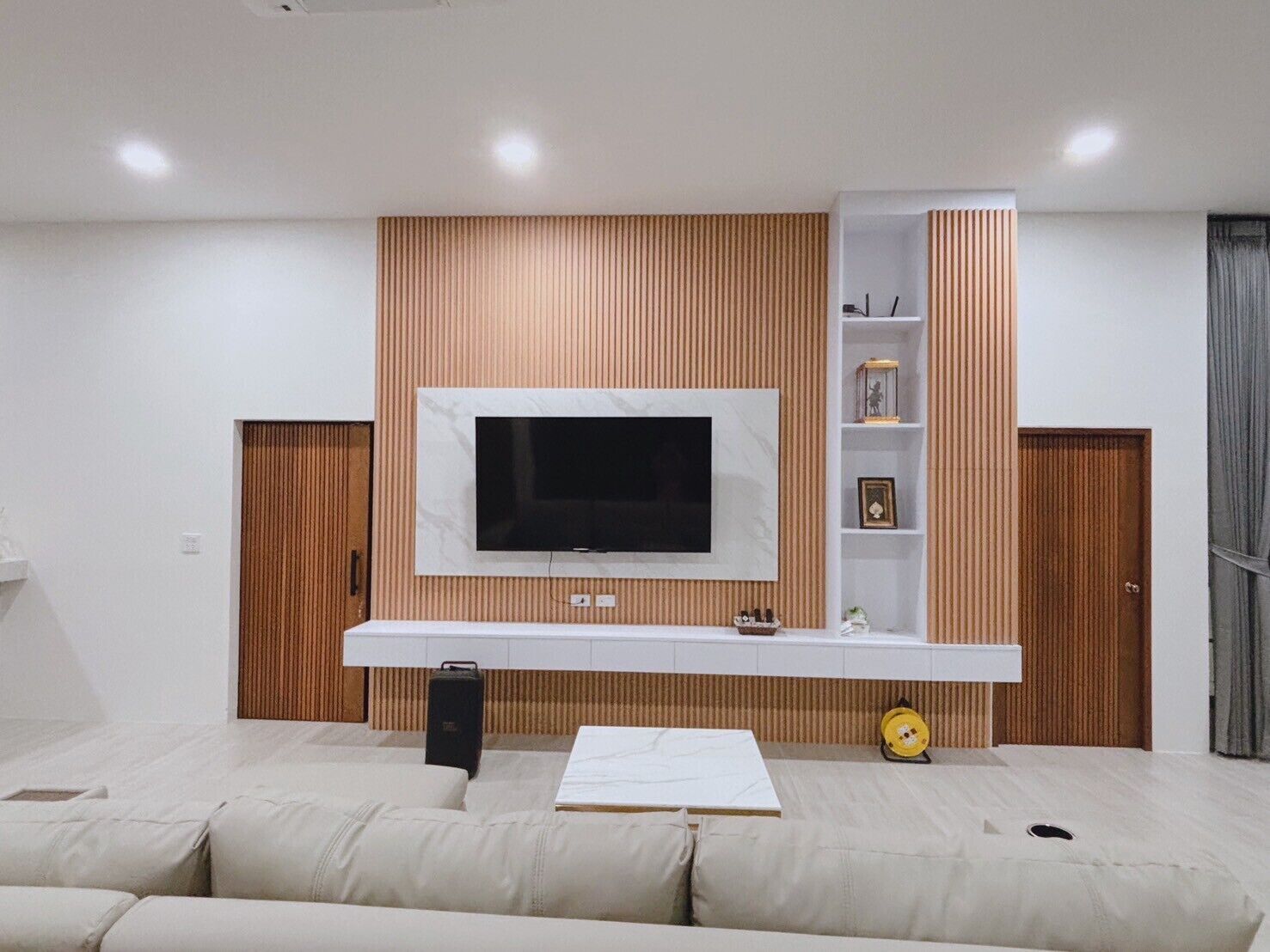 Een moderne woonkamer met een grote flatscreen televisie aan een houten wand bevestigd, een wit tv-meubel eronder en een beige bank.