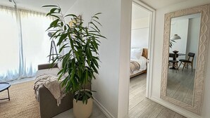 Un salon moderne avec une grande plante en pot, un canapé et un coin repas visible à travers une porte ouverte.