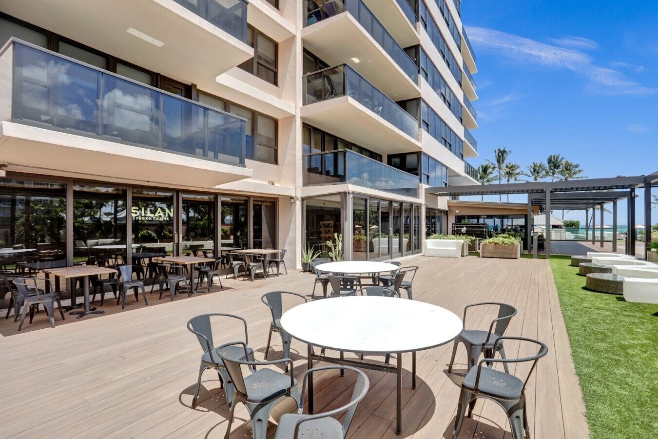 A modern outdoor dining area with multiple tables and chairs, a poolside lounge, and a restaurant with large windows.