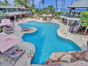 A swimming pool with lounge chairs and umbrellas, surrounded by palm trees and a view of the ocean. - Coastal Condo with Resort Service, Pools & Gym 407 (Miami Beach)