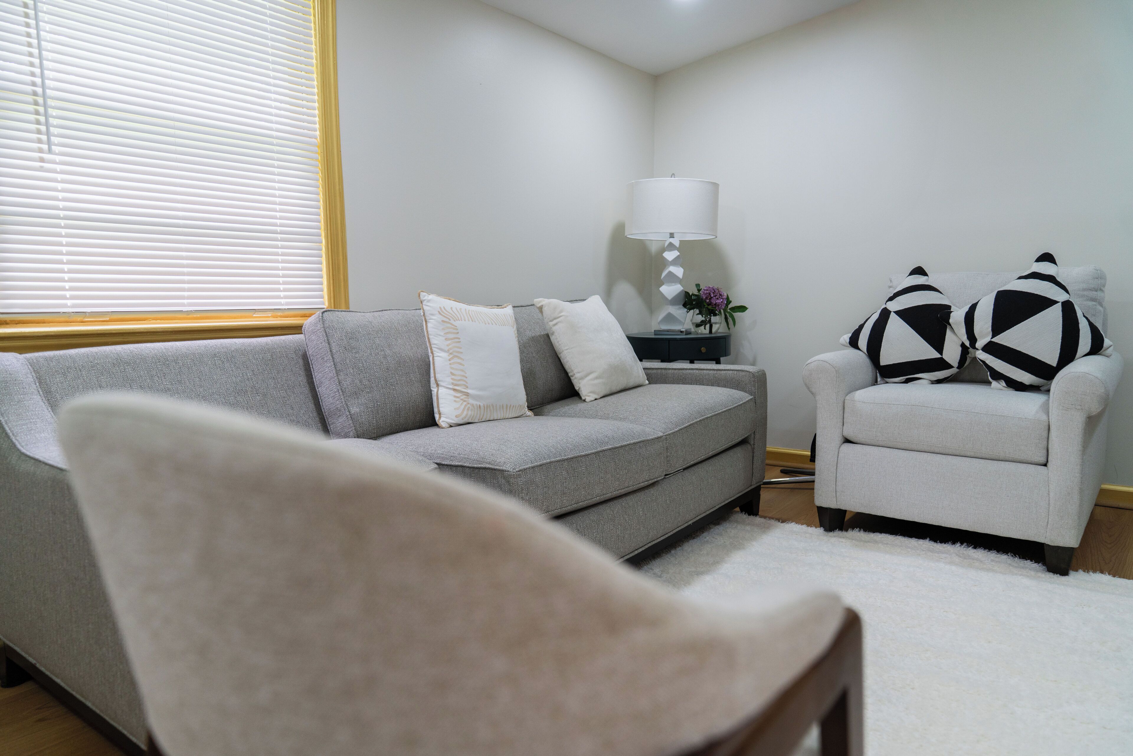 A living room with a grey sofa, a white armchair, a small side table with a lamp, and a white rug.