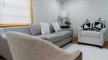 A living room with a grey sofa, a white armchair, a small side table with a lamp, and a white rug.