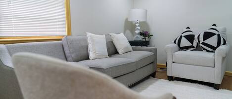 A living room with a grey sofa, a white armchair, a small side table with a lamp, and a white rug.