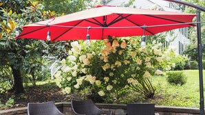 An outdoor patio with wicker furniture, a red umbrella, and a garden with white flowers. - Legacy Villa (Upper Marlboro)