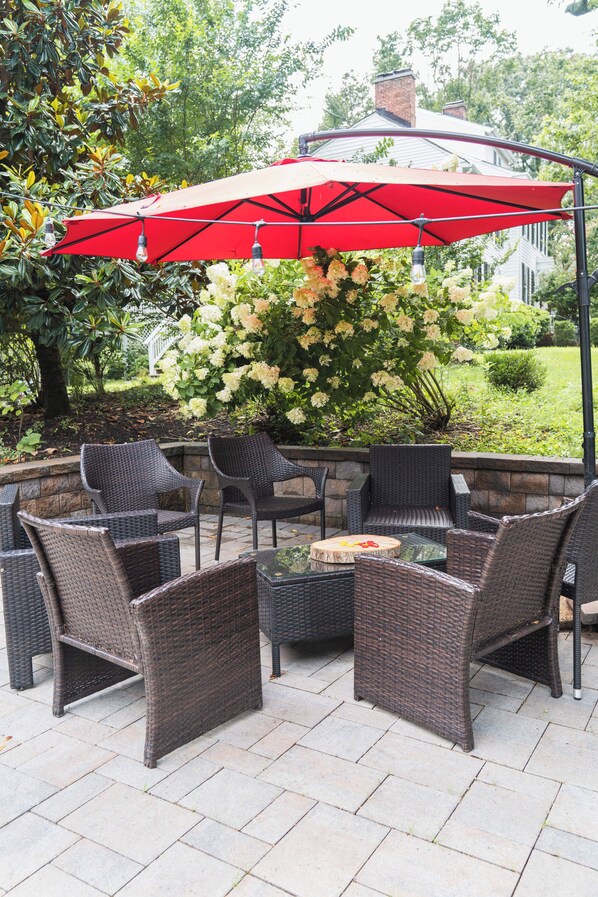 An outdoor patio with wicker furniture, a red umbrella, and a garden with white flowers. - Legacy Villa (Upper Marlboro)