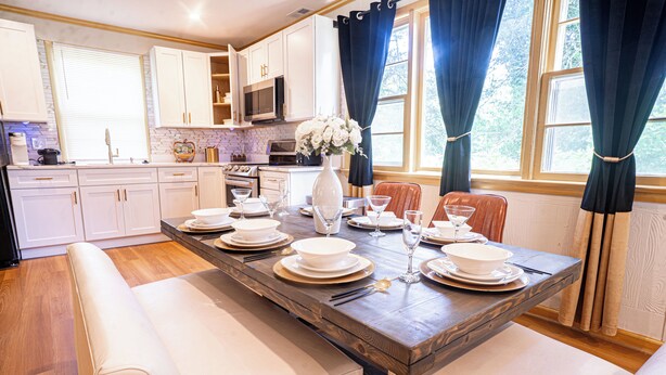 A modern kitchen with white cabinets, a wooden dining table set for four, and a view of the outdoors through large windows. - Legacy Villa (Upper Marlboro)