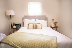 A neatly made bed with a green blanket, a bedside lamp, a small table with a plant, and a window with blinds. - Legacy Villa (Upper Marlboro)