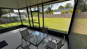 En overdækket terrasse med et glasbord og fire stole, med udsigt over en græsplæde med et træhegn.