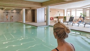 An indoor swimming pool with a clear view of the pool's edge, a wooden wall with a sign, and a seating area with lounge chairs and a person relaxing. - Apartment in France With Heated Pool & Spa (Les Belleville)
