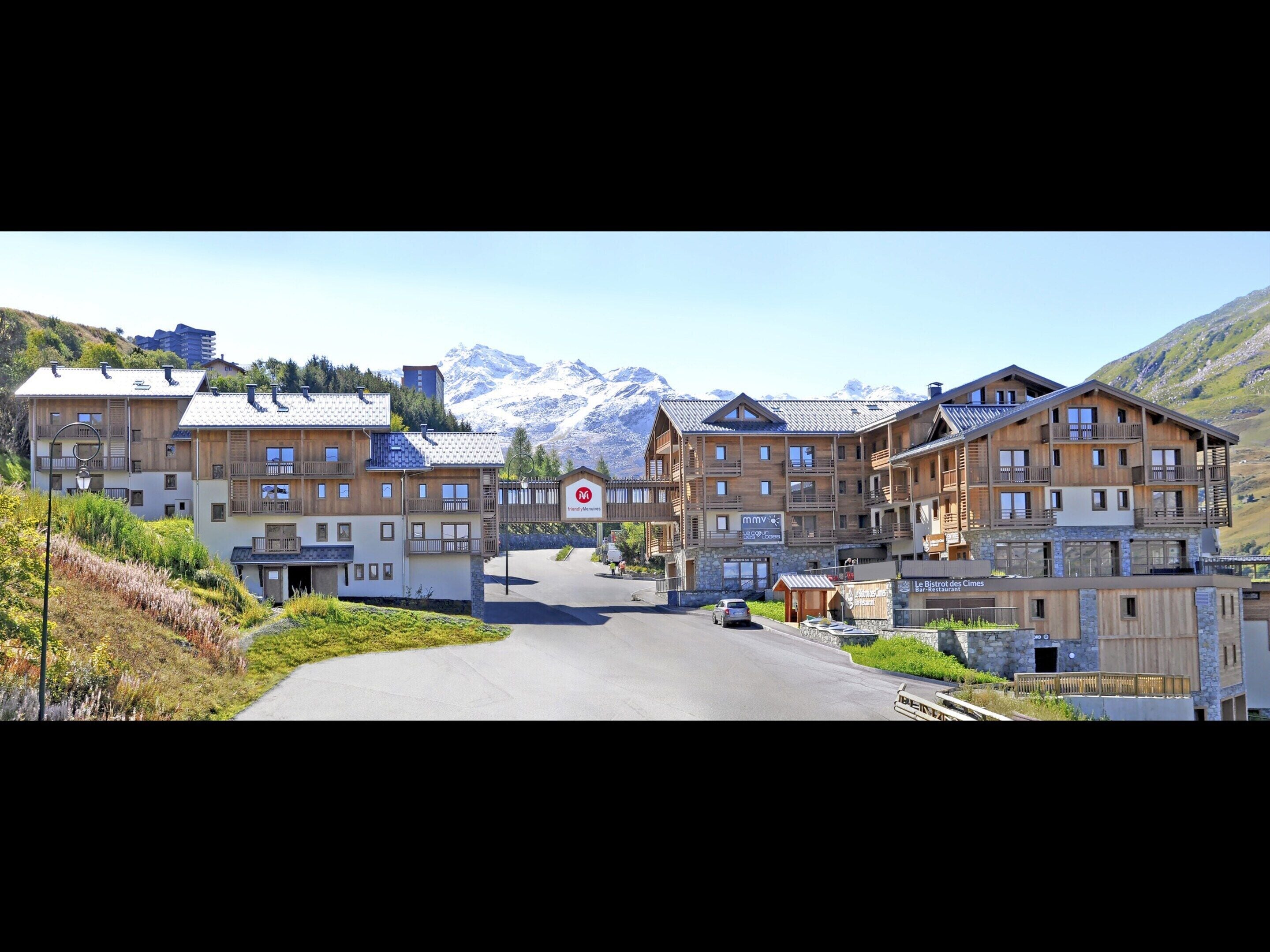 A mountain resort with wooden buildings, a road leading to the entrance, and snow-capped mountains in the background.