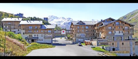 A mountain resort with wooden buildings, a road leading to the entrance, and snow-capped mountains in the background.