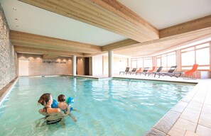 Une piscine intérieure à l’eau claire, avec des poutres en bois au plafond et des chaises longues disposées le long des bords.