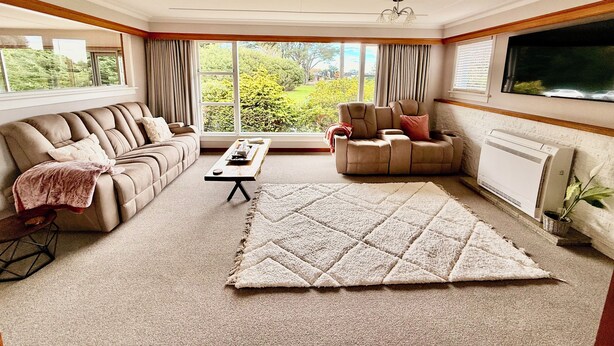 A spacious living room with a beige sofa, a recliner, a coffee table, and a large window offering a view of greenery. - Comfort at Stag Lodge Just Mins Walk to Queens Park & Down Town Invercargill (Invercargill)