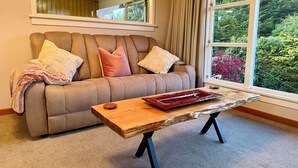 A living room with a brown sofa, a wooden coffee table, and a large window with curtains. - Comfort at Stag Lodge Just Mins Walk to Queens Park & Down Town Invercargill (Invercargill)