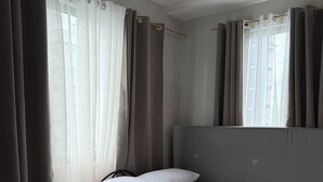 A single bed with white bedding, a grey headboard, and a window with sheer curtains.