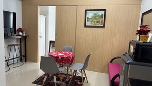 A compact living space with a dining area featuring a table with a red floral tablecloth, two grey chairs, and a small rug. A television is mounted on the wall, and a framed picture hangs above the table.