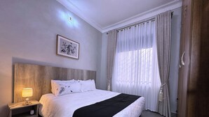 Une chambre à coucher bien rangée, avec un grand lit, une table de chevet avec une lampe, un tableau encadré au mur et une fenêtre avec des rideaux.