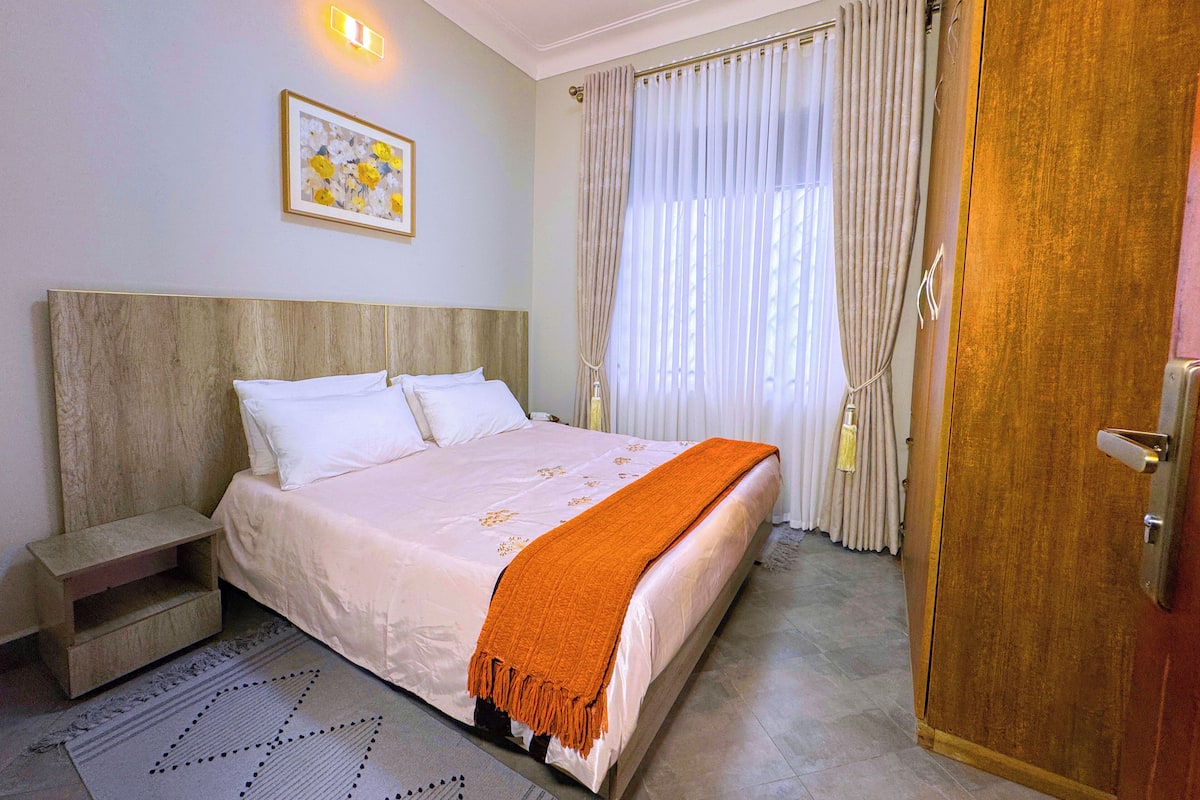 A hotel room with a large bed, a nightstand, a wooden door, and a window with curtains.