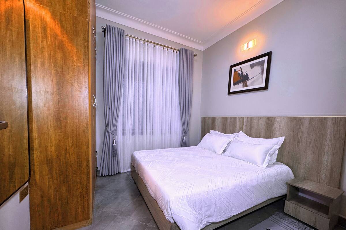 A hotel room with a large bed, a wooden headboard, a nightstand, a framed picture on the wall, and a view of the hallway and curtains.
