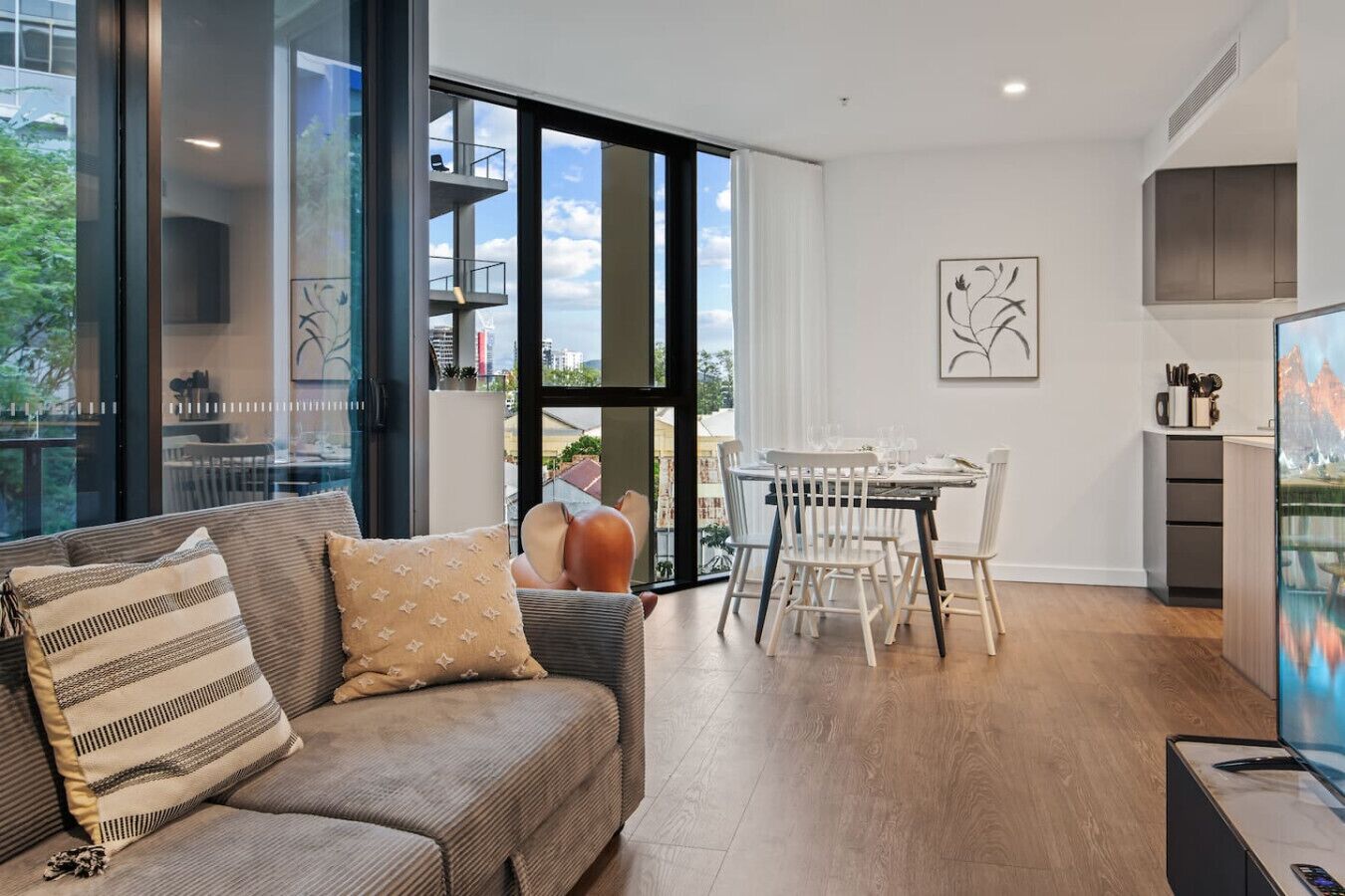 A modern living room with a sofa, dining table, and chairs. There is a large window with a view of buildings and greenery.