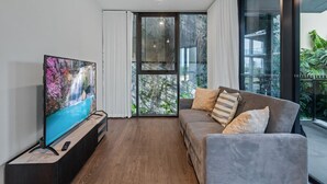 A modern living room with a flat-screen TV on a wooden stand, a grey sofa with patterned cushions, and a large sliding glass door leading to a balcony with greenery.