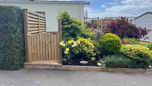 Ein von einer Hecke umgebener Gartenbereich mit verschiedenen Pflanzen und Blumen, einem hölzernen Tor und einem Gebäude im Hintergrund.