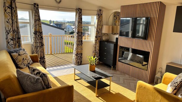 Ein Wohnzimmer mit Sofa, Sesseln, Couchtisch, Fernseher und einem Balkon mit Vorhängen.