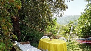 Juego de banco y mesa de madera al aire libre, con mantel amarillo, rodeado de follaje y vistas a colinas.