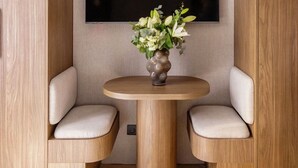 Una habitación moderna con una mesa redonda de madera, dos sillas acolchadas, un jarrón con flores y un televisor de pantalla plana montado en la pared.