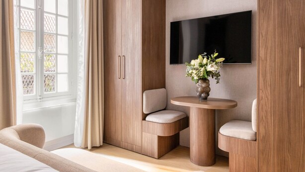Una sala de estar moderna con un televisor de pantalla plana montado en la pared, una mesa redonda de madera con dos sillas y un amplio ventanal con cortinas.
