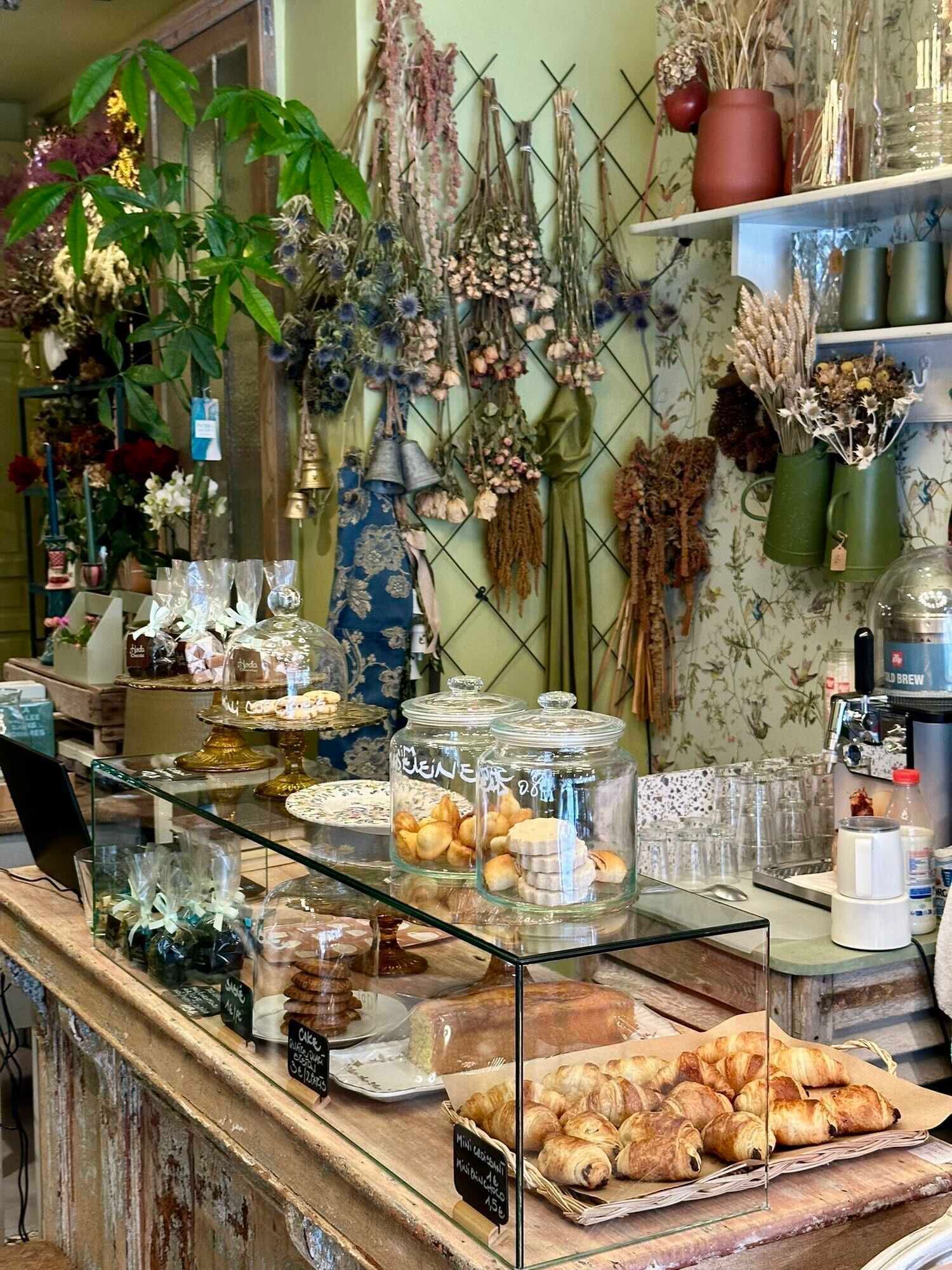 Bancada de pastelaria com doces, uma máquina de café e vários flores e plantas ao fundo.