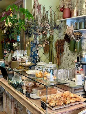A bakery counter with pastries, a coffee machine, and various flowers and plants in the background.