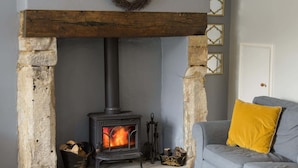 Une cheminée en pierre avec un poêle noir, une cheminée en bois et un canapé gris agrémenté d’un coussin jaune.