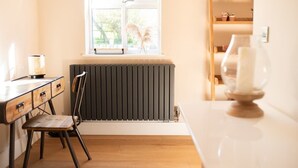 A room with a radiator, a window with a view of greenery, a desk with drawers, a chair, and a bedside table with a candle. - Vineyard Style 2BR with Garden and Parking (Shudy Camps)