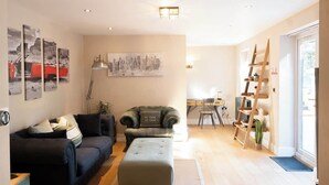 A living room with a sofa, ottoman, and a painting of a cityscape. There is a ladder against the wall and a desk with a chair. - Vineyard Style 2BR with Garden and Parking (Shudy Camps)