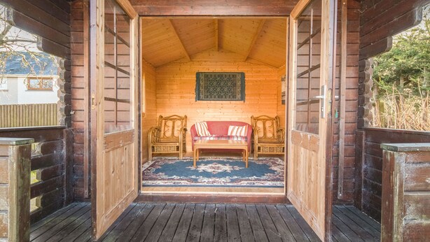 A wooden cabin interior with a red sofa, two wooden chairs, and a patterned rug.