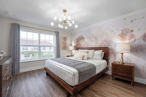 Une chambre avec un grand lit, un plancher en bois, une fenêtre avec des rideaux, une table de chevet avec une lampe et un mur orné d’œuvres d’art abstraites.