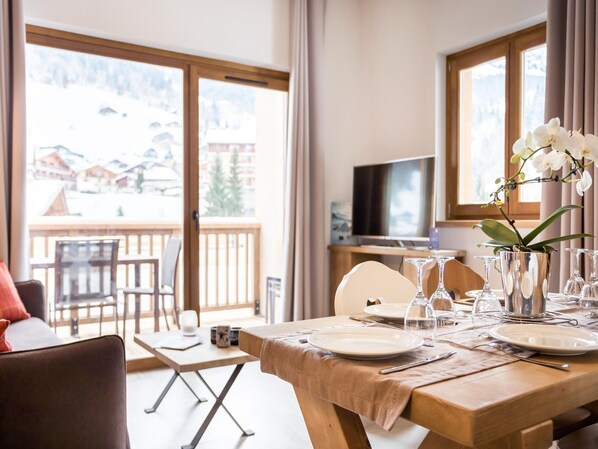 Un coin repas avec une table dressée pour deux personnes, un téléviseur en arrière-plan et un balcon offrant une vue sur les bâtiments et les arbres.