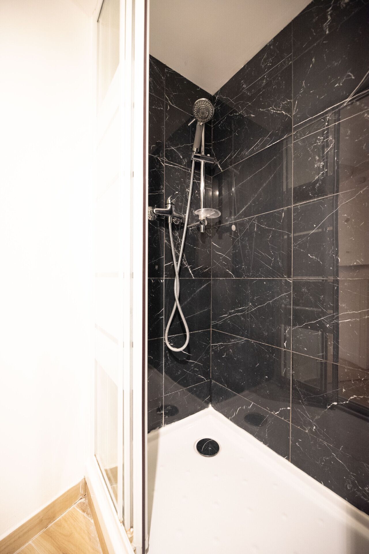 Un baño moderno con paredes de mármol negro, una ducha con cabezal de ducha manual y un lavamanos blanco.