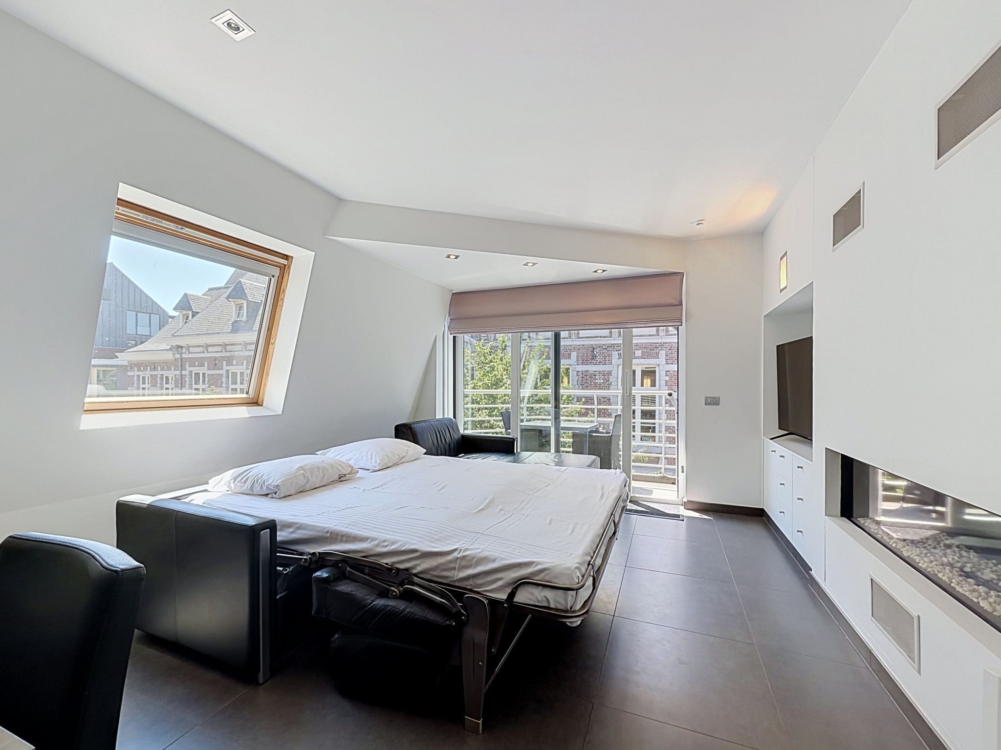 A modern bedroom with a bed, a desk, a chair, a TV, and a balcony with a view of buildings.