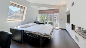 Un dormitorio moderno con cama, escritorio, silla, televisor y balcón con vista a edificios.