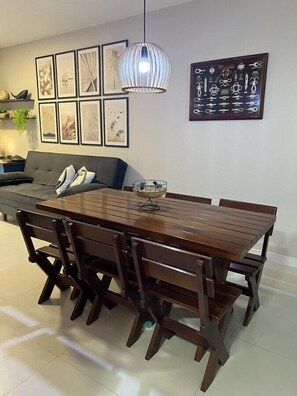 A dining area with a wooden table and chairs, a hanging light fixture, and framed artwork on the walls. - Pé na Areia Itapema · 3 Quartos · Churrasqueira (Itapema)