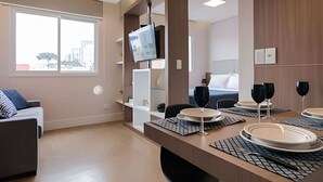 A modern hotel room with a large bed, a TV mounted on a wooden panel, a dining table set with plates and glasses, and a large window offering a city view. - Studio prox shopping Curitiba Wifi, TV a Cabo e ar (Curitiba)