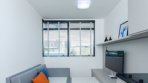 Una sala moderna con un sofá gris, un televisor de pantalla plana y un ventanal con persianas.