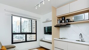 Une cuisine moderne avec des meubles blancs, un four à micro-ondes et une machine à café. Une grande fenêtre offre une vue sur les bâtiments environnants.