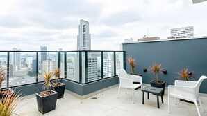 Une terrasse sur le toit, dotée d’une balustrade en verre, de plantes en pot, de chaises blanches et offrant une vue sur les bâtiments de la ville.