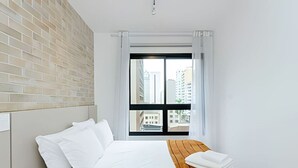 Une chambre d’hôtel avec un lit, une tête de lit en bois, une fenêtre avec des rideaux et une vue sur les bâtiments environnants.