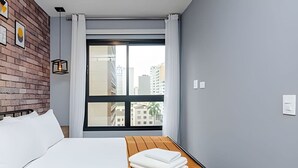 A hotel room with a bed, a wooden headboard, a large window with a view of buildings, and a brick wall with framed pictures.
