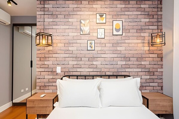 Une chambre avec un mur en briques, un lit avec une literie blanche, deux tables de chevet et des lampes fixées au mur.