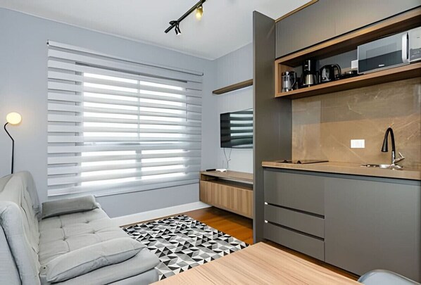 A modern kitchen with a built-in microwave, a coffee maker, and a sink. There is a grey sofa and a patterned rug in the living area.